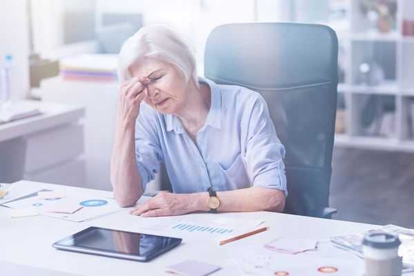 Older woman at a desk holding her forehead, distressed over finances.